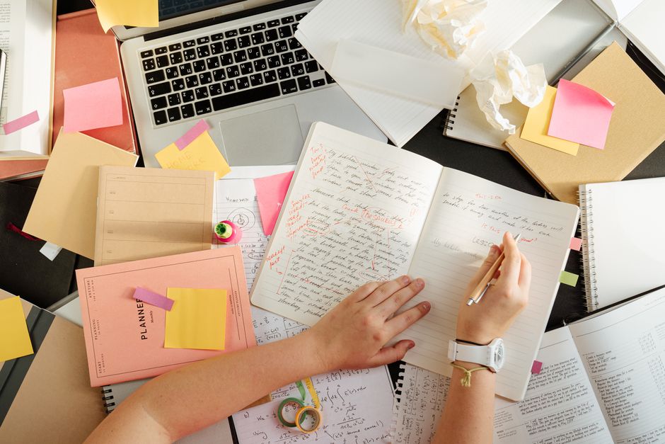 Overhead view of a study desk with a laptop, planner, sticky notes, and a hand writing in a notebook