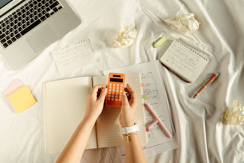 Hands using a calculator over an open notebook with math notes and a laptop